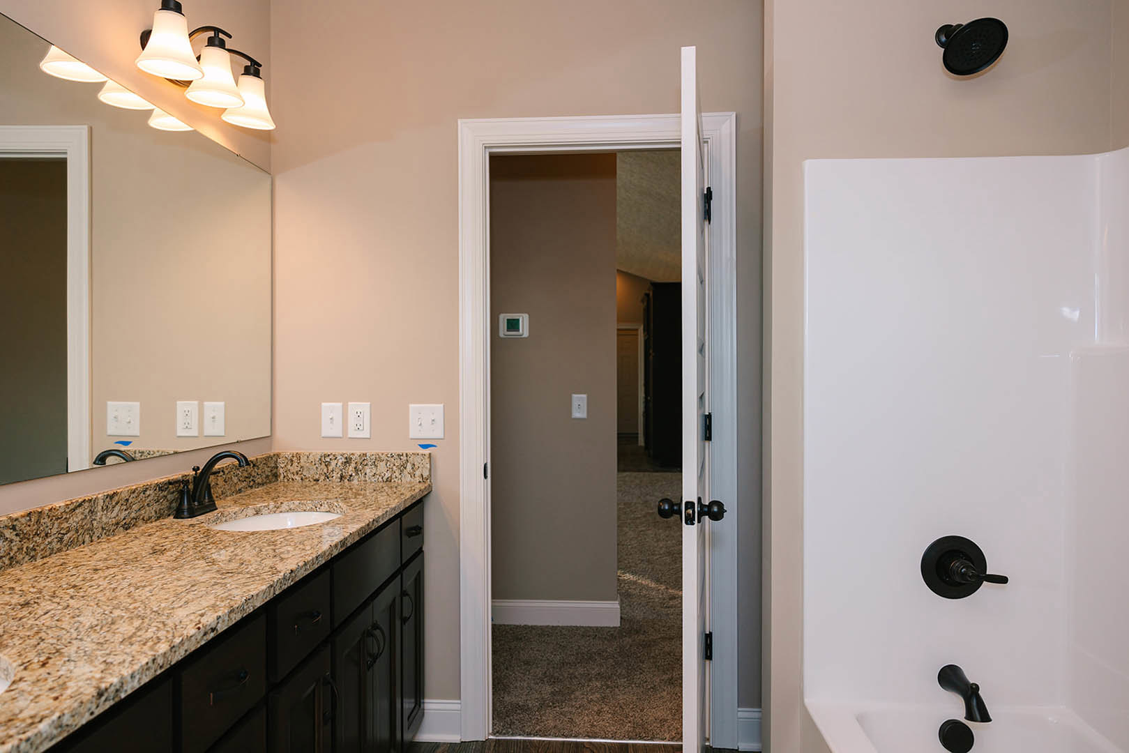 White bathroom with rectangular mirror above a stone countertop, undermount sink, black faucet, white tile backsplash, black shower head and handle, white wall with black knob and