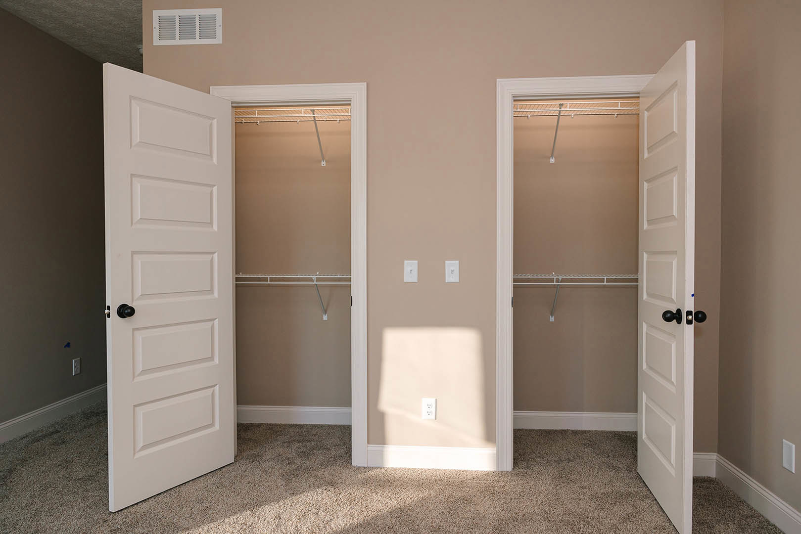 Walk-in closet with open white doors, built-in white shelving, carpeted floor, and wall vent