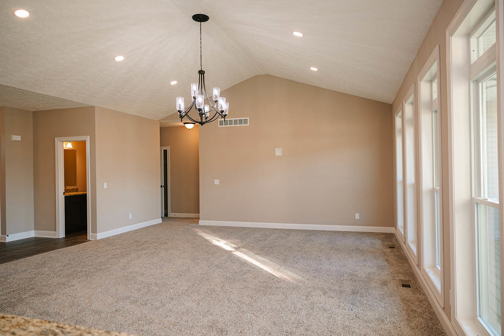 Carpeted room with sunlight streaming in, clear glass chandelier hanging from ceiling, white walls with crown molding, doorway leading to bathroom