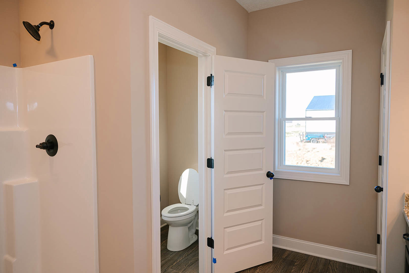White toilet with lid up beside a window, blue truck visible outside; close-up of chrome shower head, black metal fixture with round base, white door featuring black knobs
