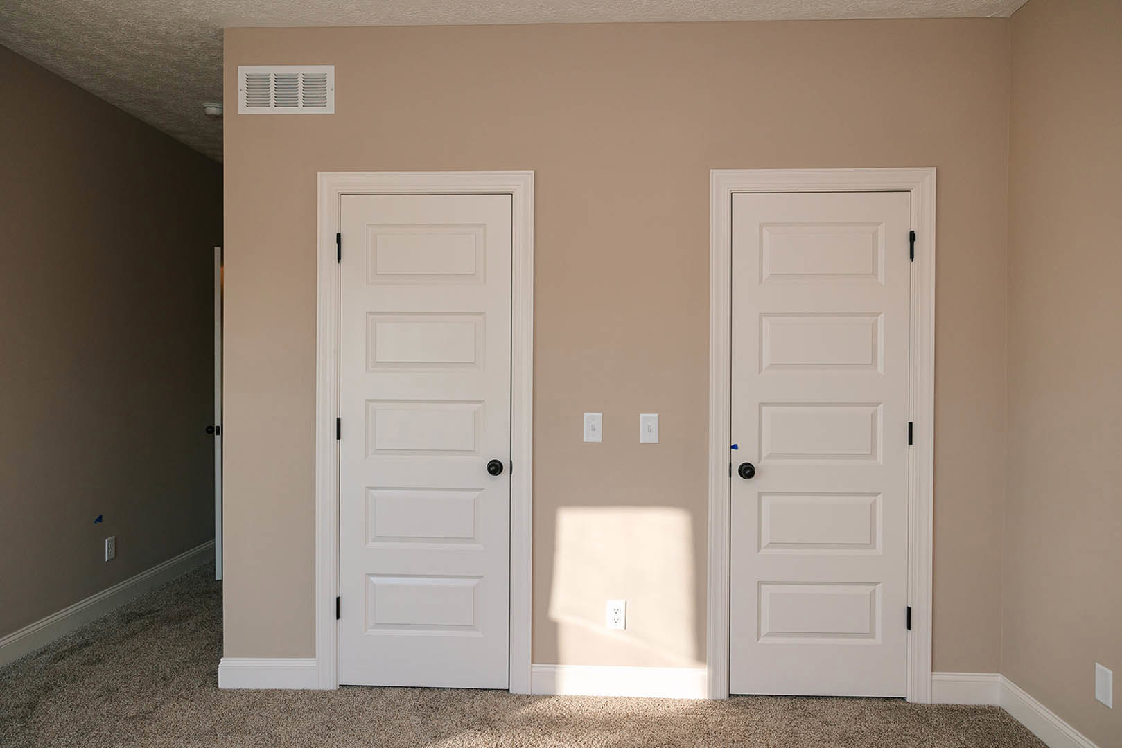 Two white paneled doors with black knobs set in a dark wall, white trim, carpeted floor, visible vent and electrical outlet