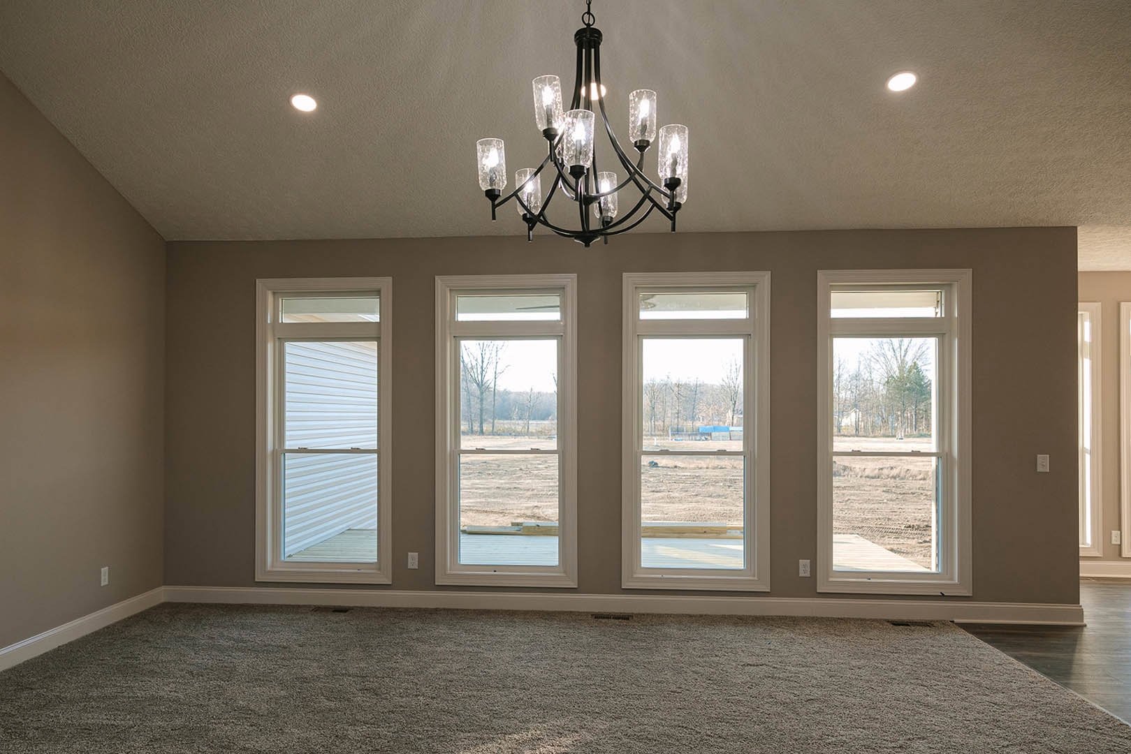 Chandelier with clear glass shades hangs from a white ceiling with crown molding, large windows with white panels overlook a grassy field, neutral carpet flooring