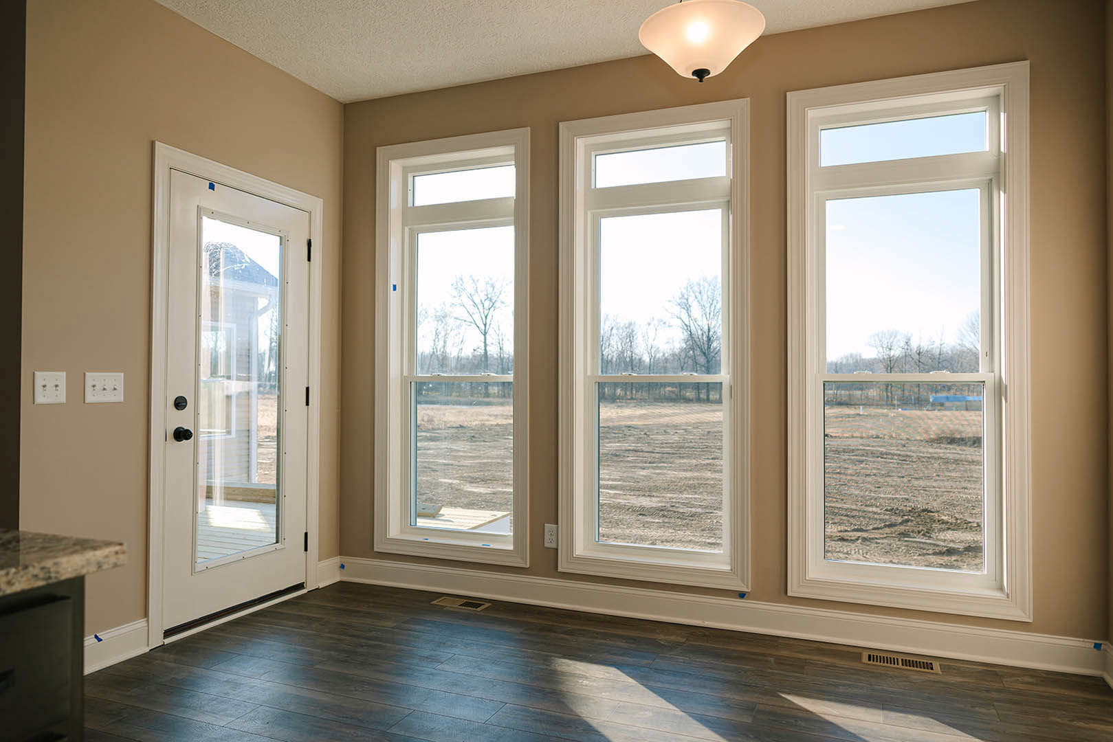 Spacious room featuring large windows with views of trees and a field, glass door, light fixture, neutral walls, and hardwood flooring
