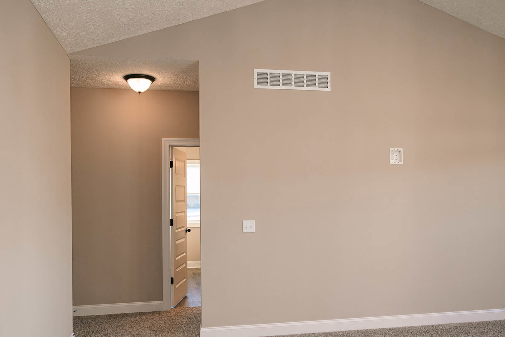 Open white door leading into a room with light-colored plaster walls, wood flooring, ceiling-mounted light fixture, white electrical outlet and light switch, and wall vent.