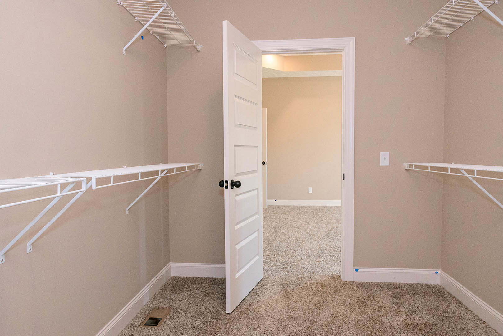White paneled door with black knobs open to a carpeted room, built-in white shelving on wall, floor vent visible near baseboard.