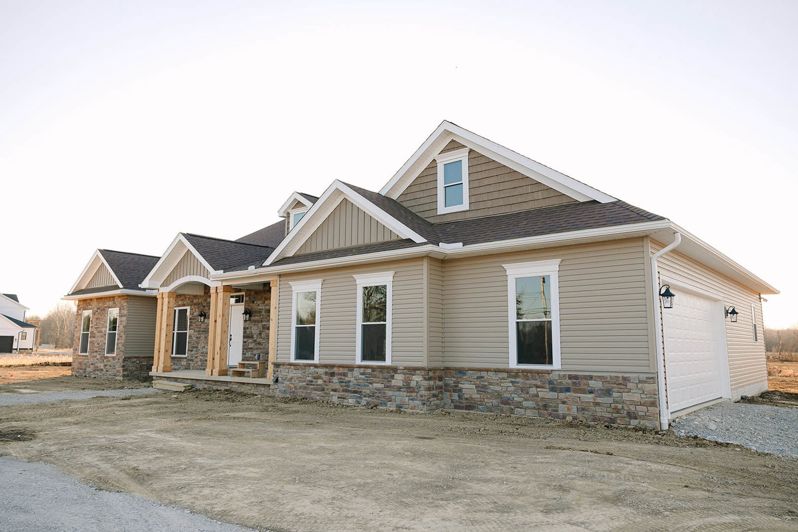 Partially built house with stone and brick exterior walls, white-framed windows, dirt driveway, and visible roof structure under clear sky