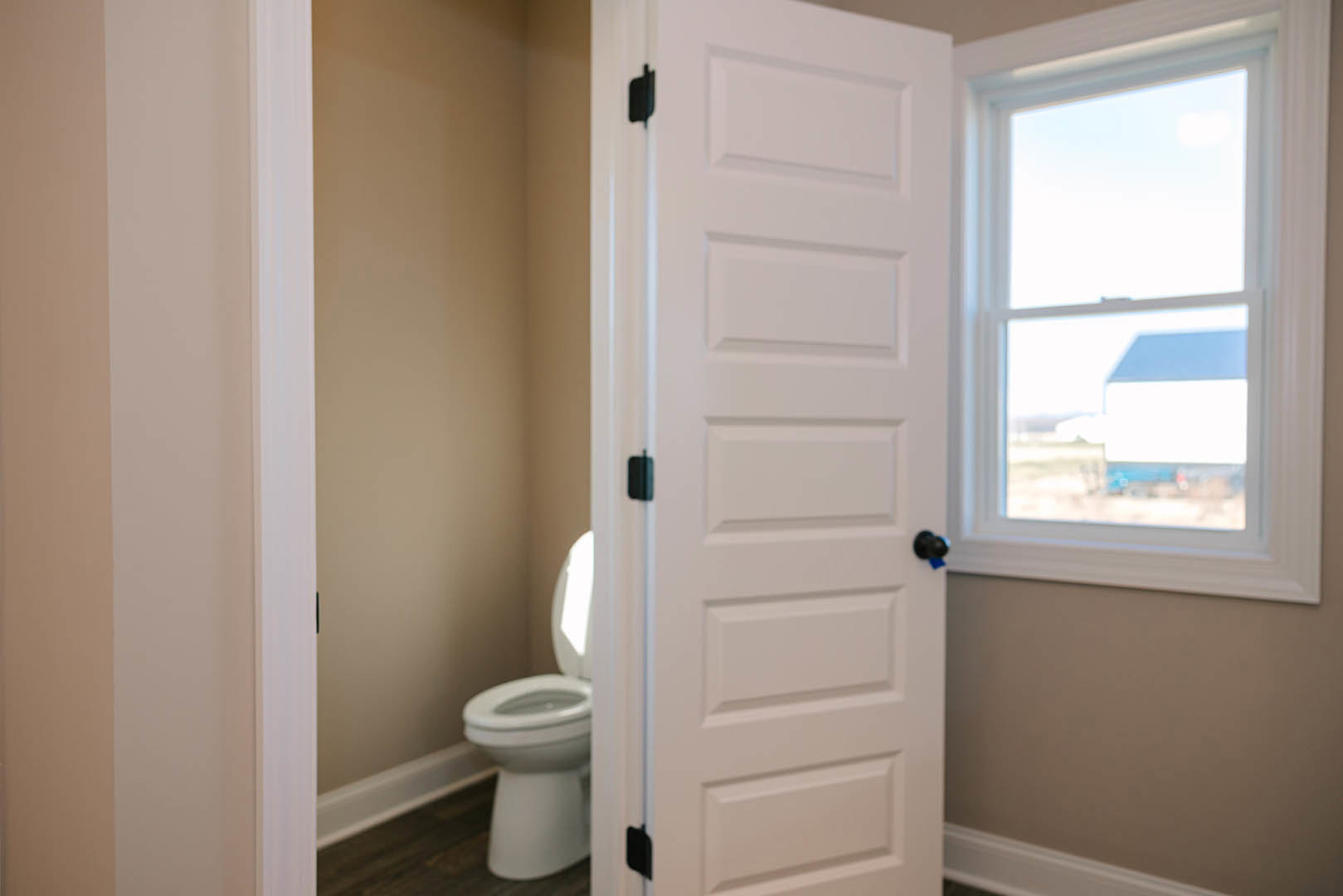 White bathroom door with black handle open to reveal toilet, window with exterior house view, light-colored walls, and minimal decor.