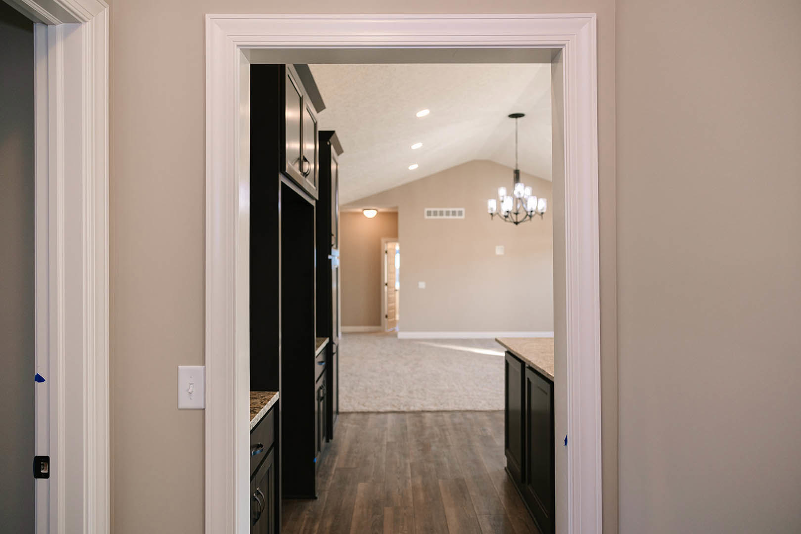 Wood floor and white wall with doorway, light switch, and chandelier; carpeted area visible in foreground, ceiling molding and mirror in background.