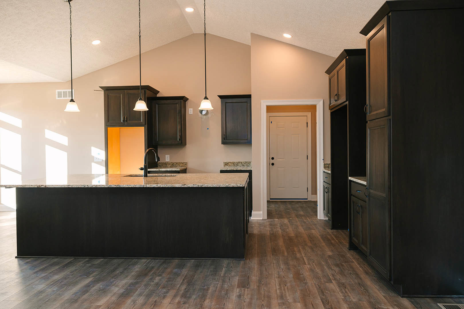 Kitchen with dark wood cabinets, marble countertops, wood flooring, white door with black knobs, bell-shaped light fixture, and stainless steel sink.