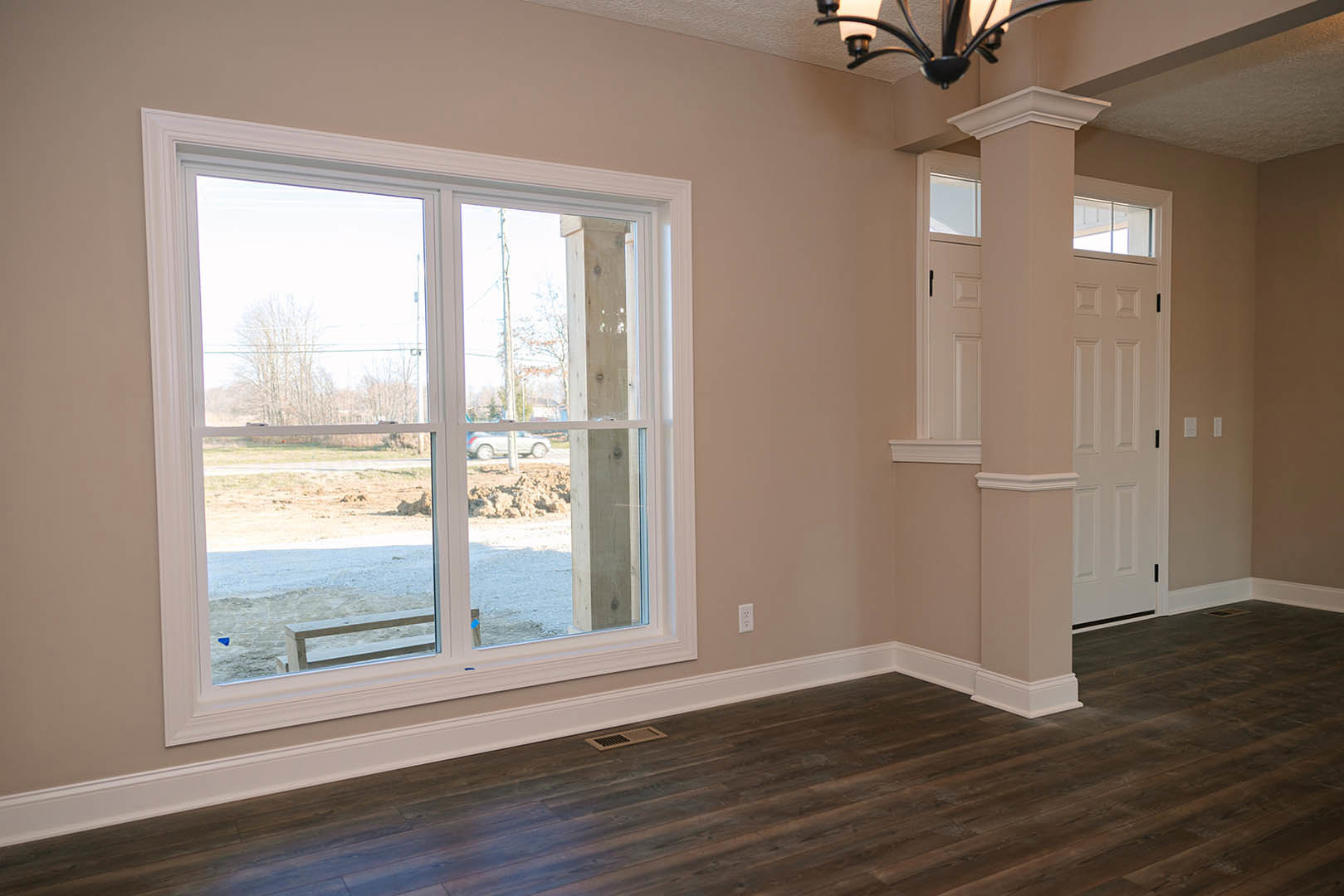 Sunlit room featuring wide wood laminate flooring, large window overlooking dirt road and trees, white walls with crown molding, ceiling light fixture, floor vent, and metal stair
