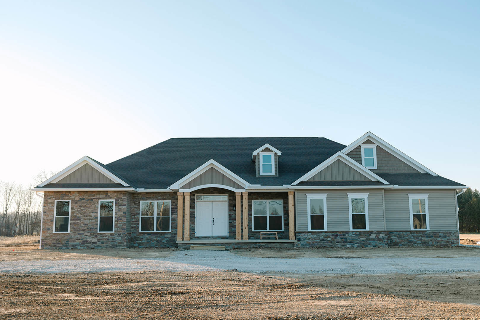 Two-story home with white siding, double front doors with glass panels, covered porch, large windows with white frames, bare dirt yard, and trees in the background
