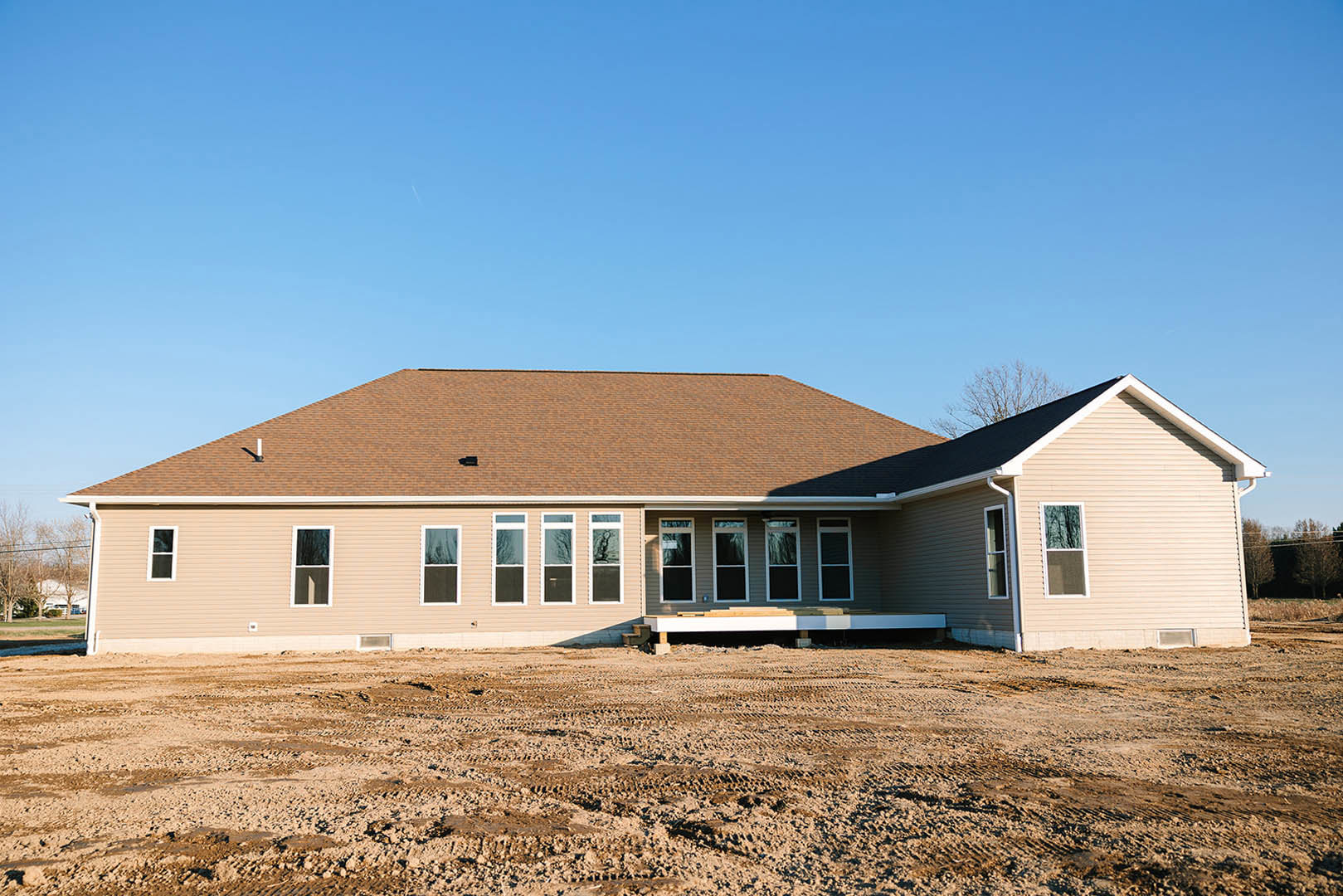 Partially built house with exposed wooden deck, white-framed window, dirt lot in foreground, and blue sky overhead