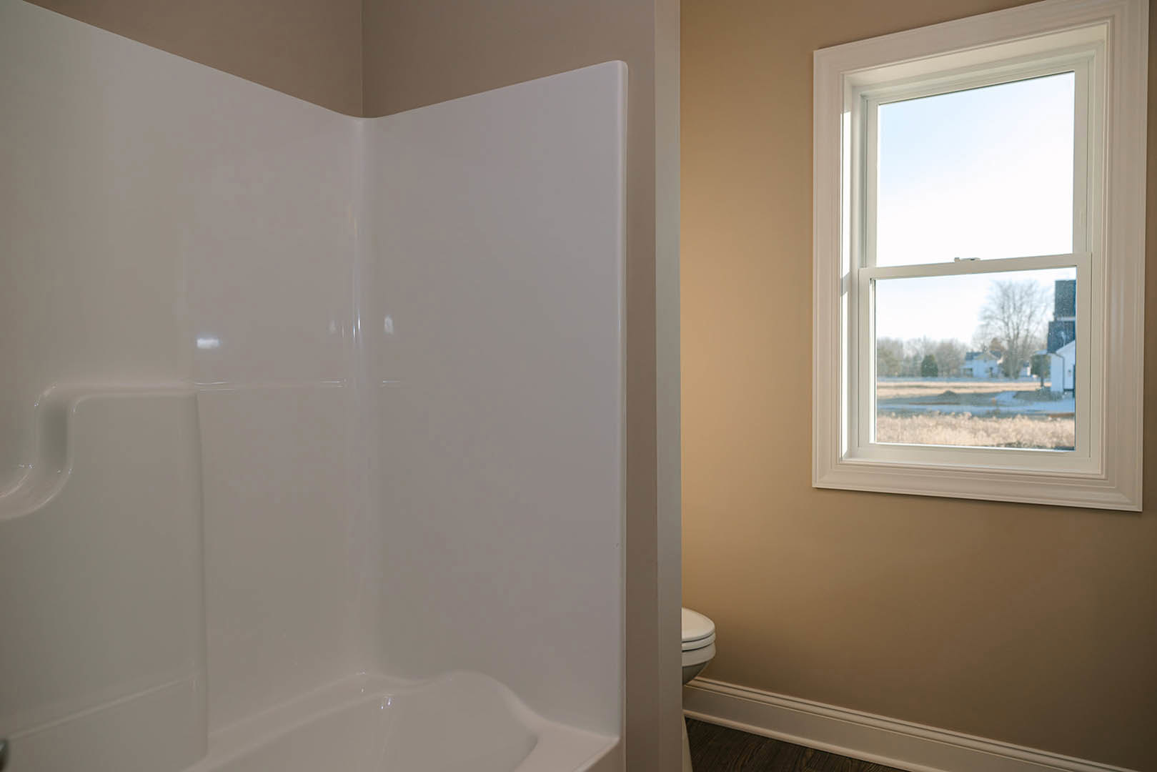 Bathroom with freestanding white tub beneath a window overlooking a field and houses, white shower stall, toilet, brown tile floor, and white walls