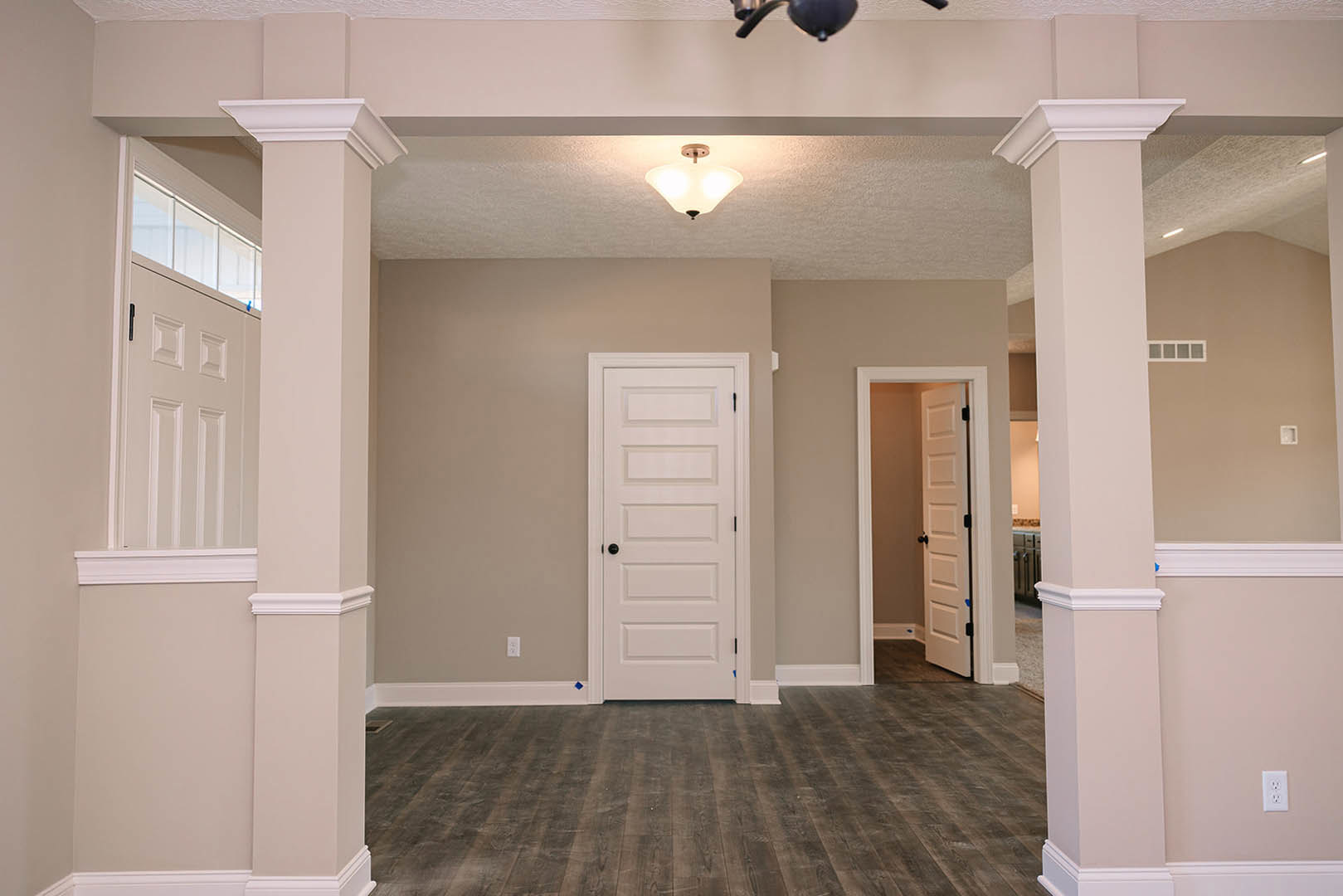 Room with white columns, white paneled door featuring black knob, wood flooring, ceiling fan, and decorative molding
