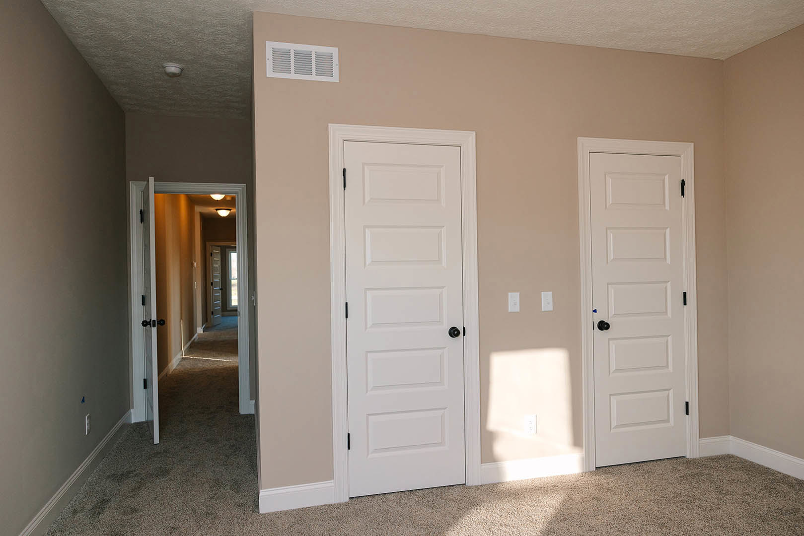 Beige-walled room featuring two white doors with black knobs, white wall vent, and hallway with ceiling light fixture