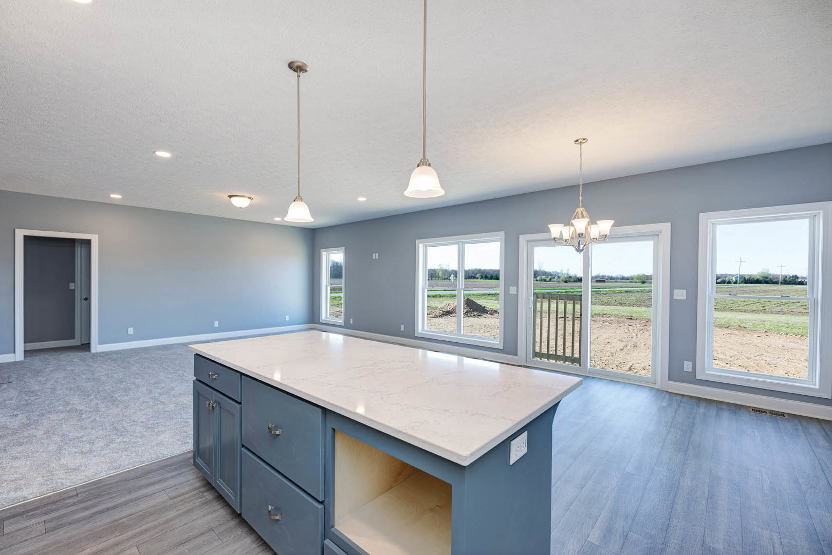 Spacious kitchen featuring a large central island with a built-in shelf, white countertops, cabinetry, a white electrical outlet, window overlooking a field, grey door with white
