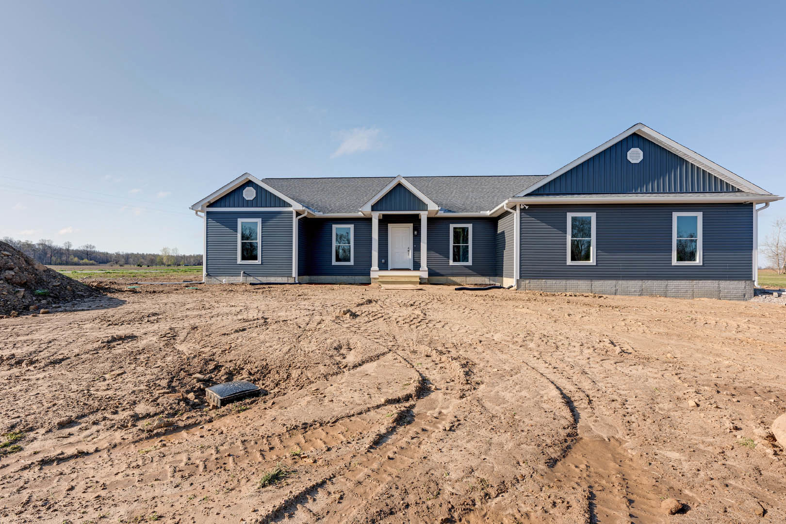 Modern house with white siding, black trim, white front door, and large window; dirt yard with visible tire tracks and a black object near the entrance