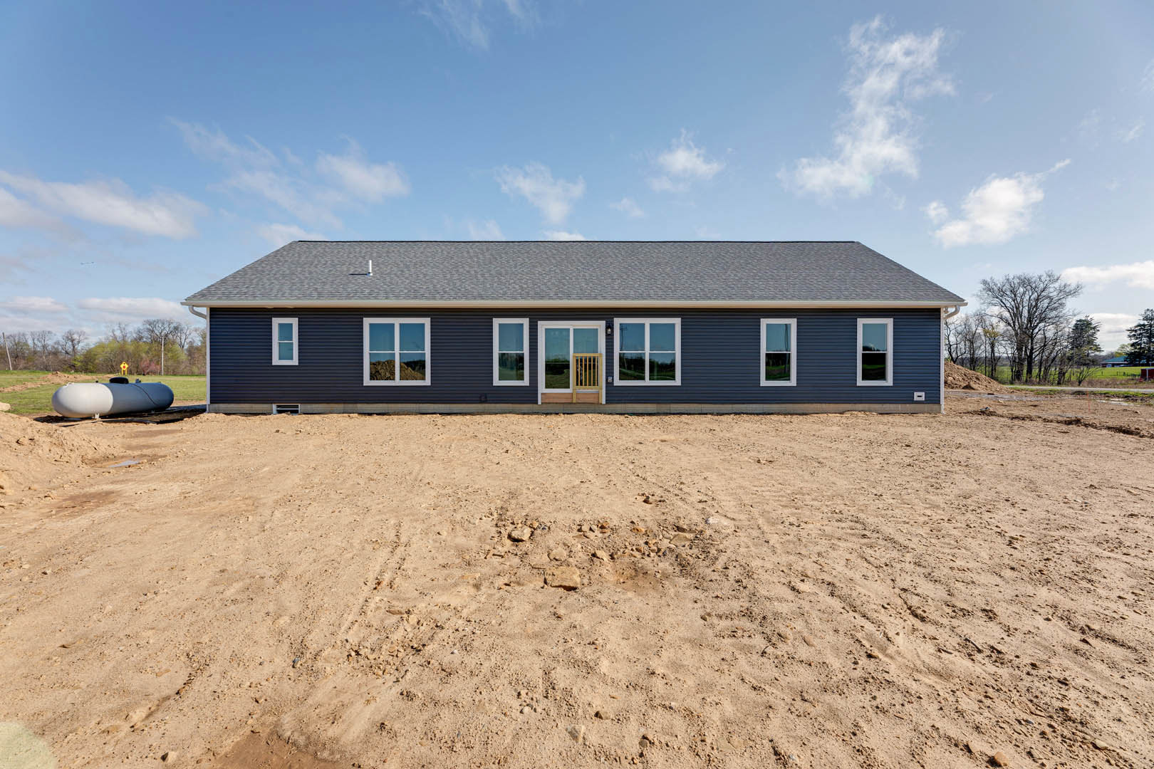 Single-story house with open door, white-framed window, and black-and-white tank, set on a dirt field with sparse trees under a cloudy sky