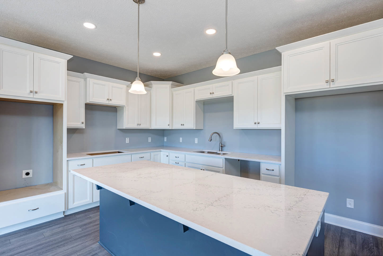 White kitchen with shaker cabinets, large central island featuring quartz countertop, stainless steel faucet, pendant light fixtures, tile backsplash, and hardwood flooring.