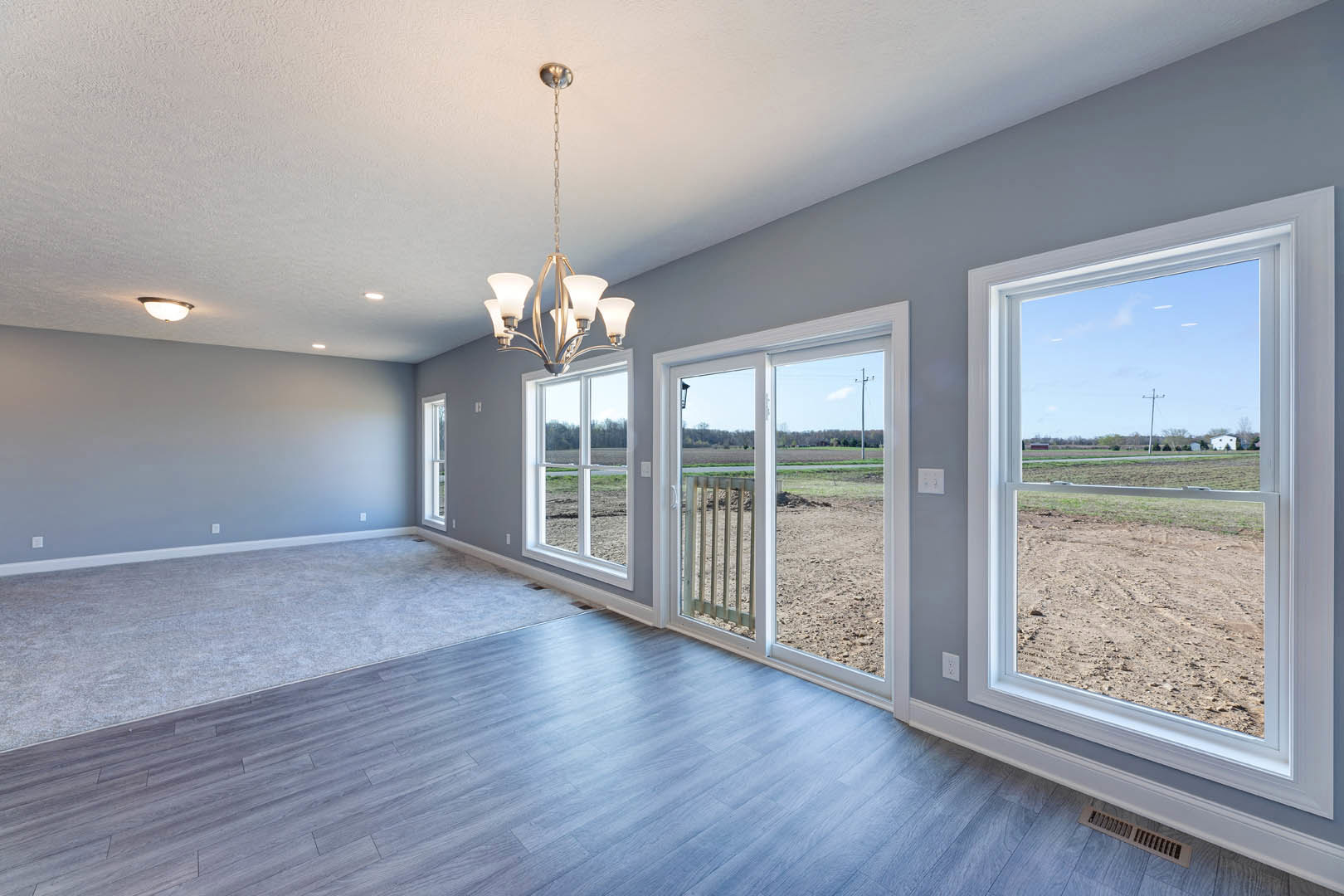 Bright room with large windows, wood flooring, ceiling chandelier, floor vent, and sliding glass door overlooking an open field