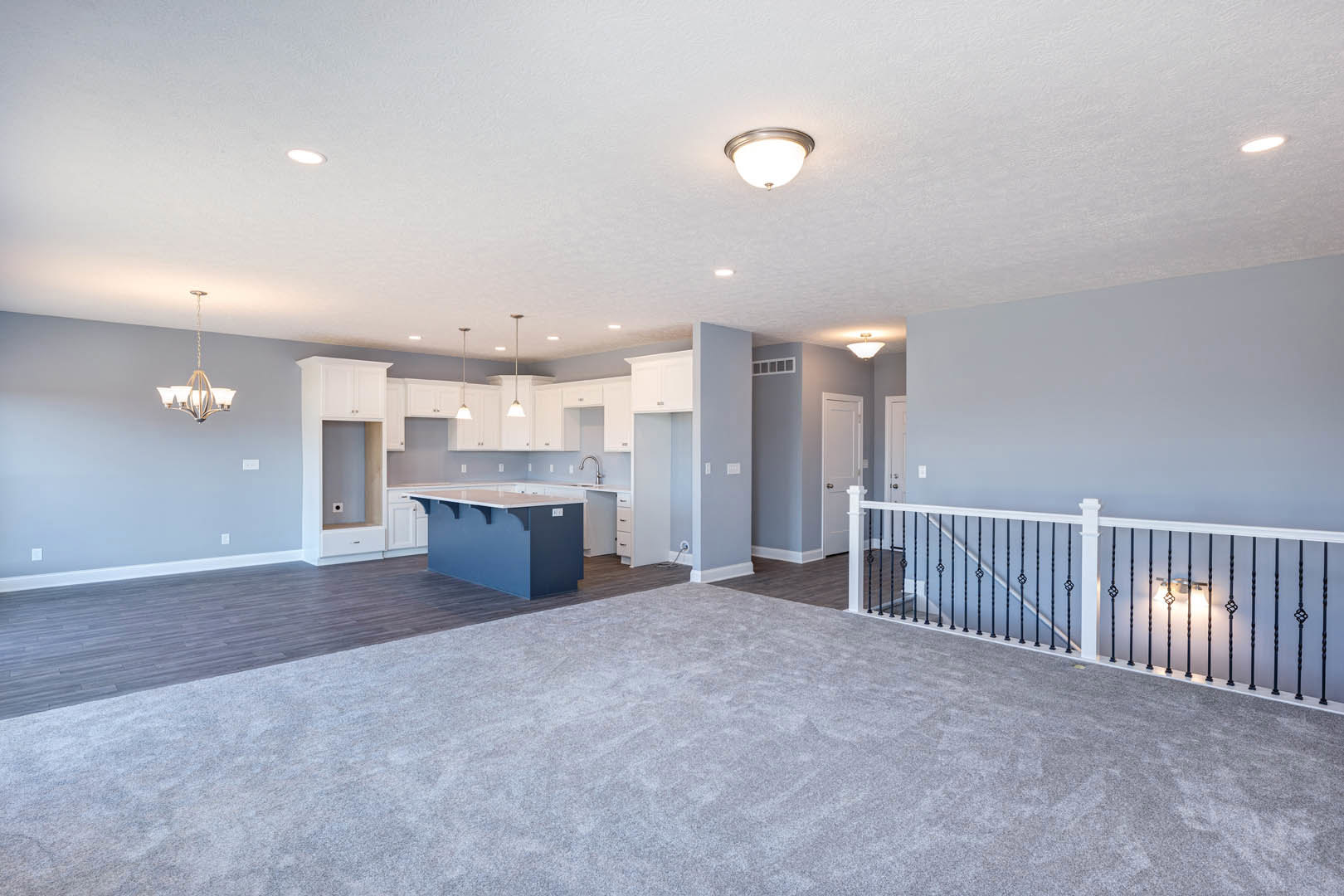 Open-concept living area featuring a kitchen with blue countertops, white cabinetry, and pendant lighting; adjacent staircase with white and black railings; carpeted section and