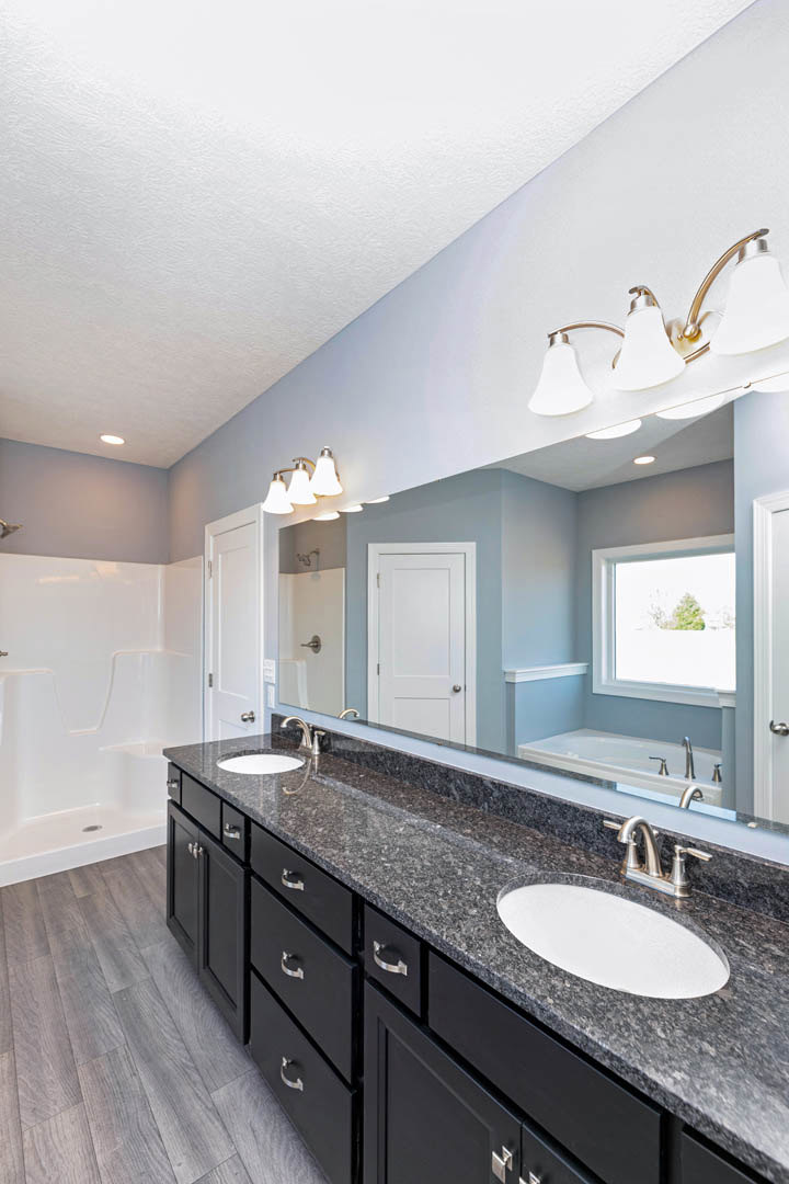Bathroom with a white oval sink on a marble countertop, large mirror above, white door with silver knobs, close-up of white tiled shower, window showing trees, and four-lamp light