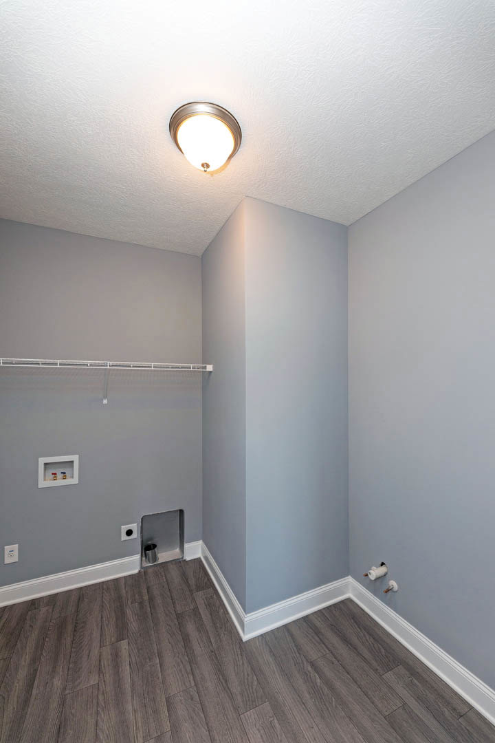 Open room with warm wood flooring, white walls, ceiling-mounted light fixture, white baseboard trim, and metal wall rod.