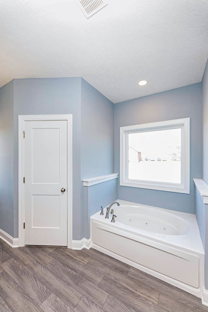White bathtub with silver faucets beside a window with a white frame, wood flooring, white door with silver handle, and floor vent visible.