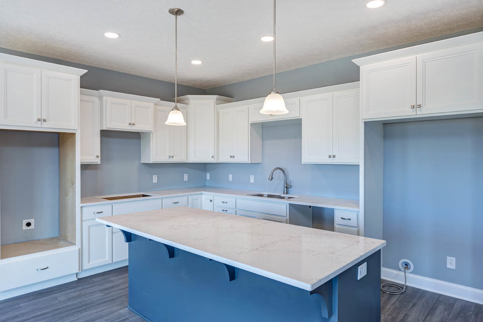 White kitchen with shaker cabinets, large central island with white countertop, stainless steel sink and faucet, pendant lights above island, light wood flooring, smooth white