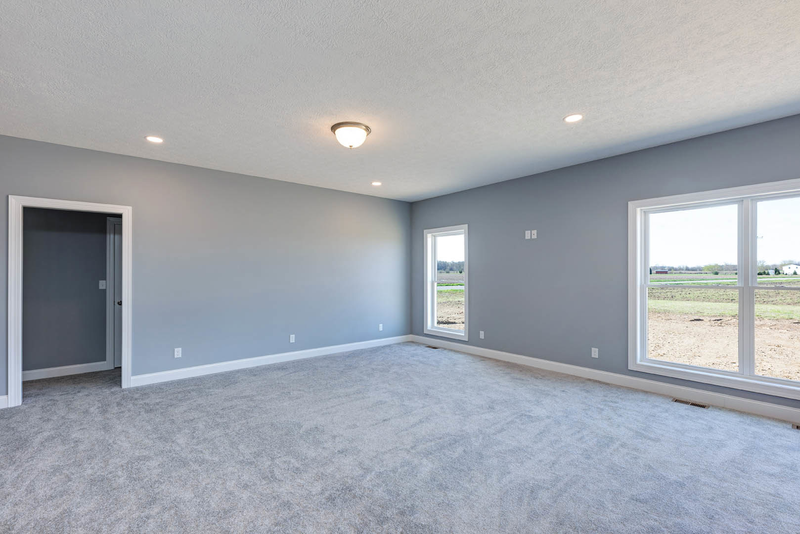 Carpeted room with large window overlooking a field, white-framed door, neutral walls, and ceiling-mounted light fixture