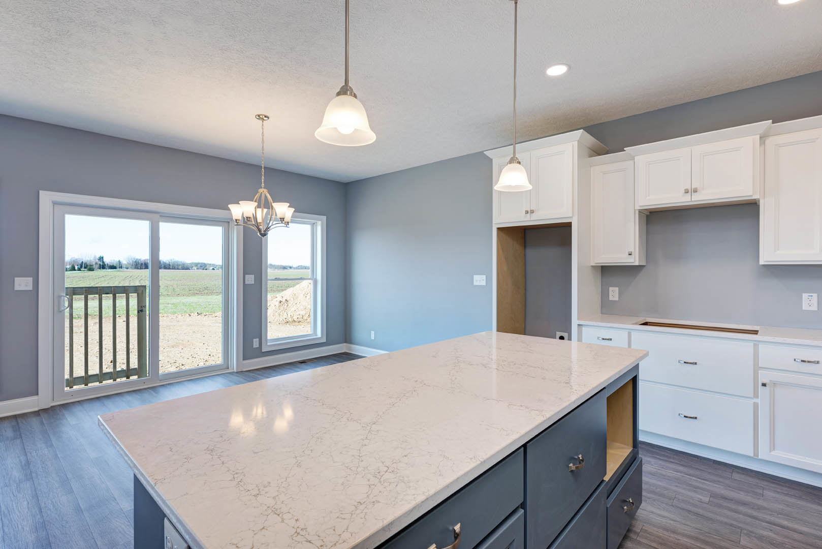 Spacious kitchen featuring a large white marble island, modern cabinetry, stainless steel sink, globe pendant light, sliding glass door opening to a field, and light wood flooring.