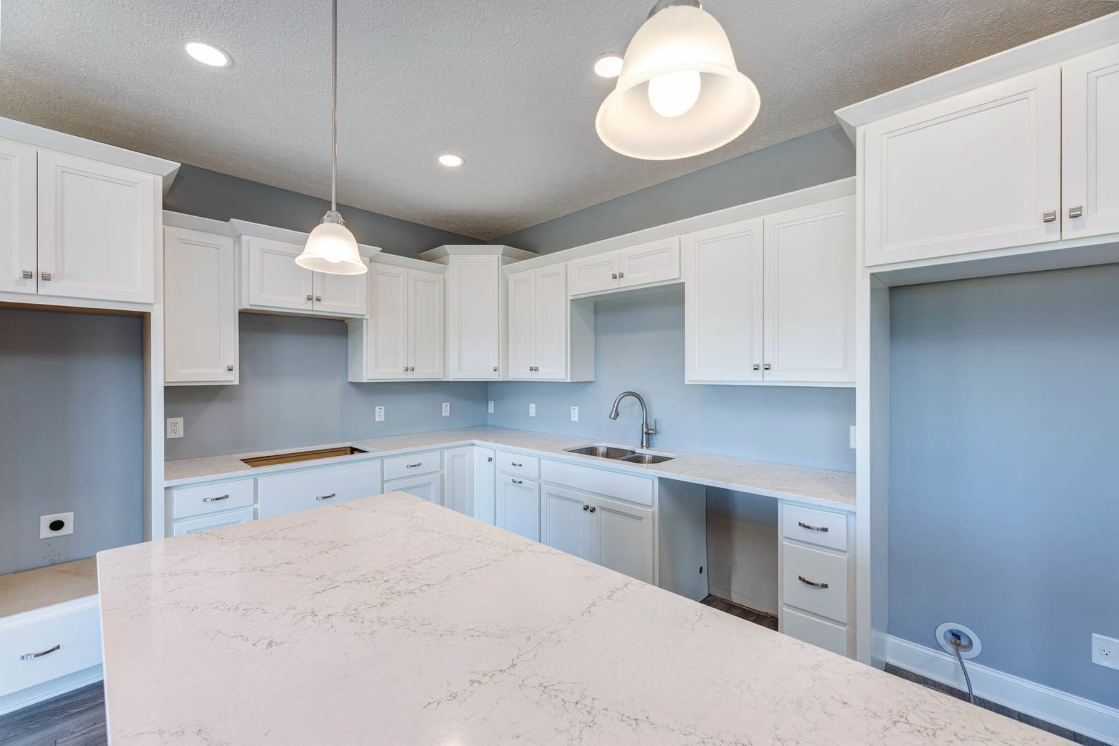 White kitchen with marble countertop, stainless steel faucet, pendant light fixture, blue accent wall with white trim, and upper and lower cabinetry.