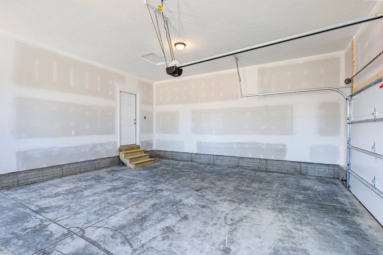White plaster walls, white door with silver handles, concrete floor with tile finish, wooden staircase beside a large window, ceiling detail visible.