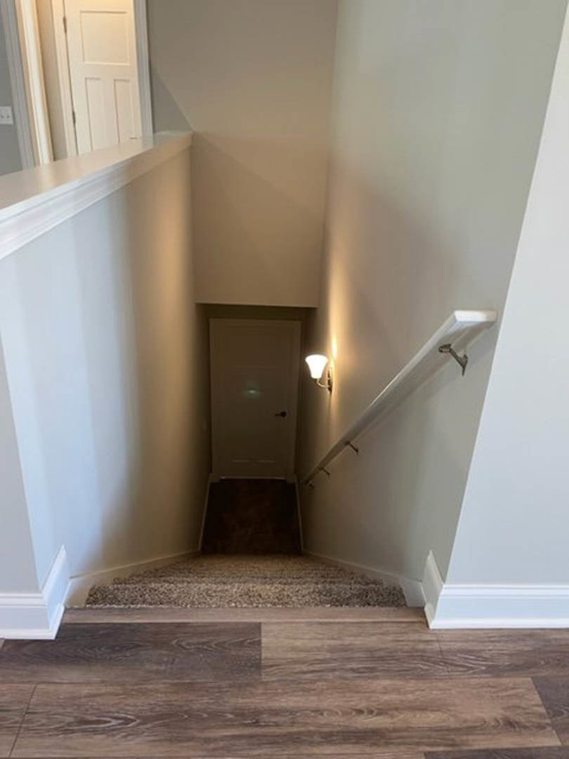 Wood staircase with white walls, white door at landing, wall-mounted sconce light, dark carpeted floor adjacent to wood flooring