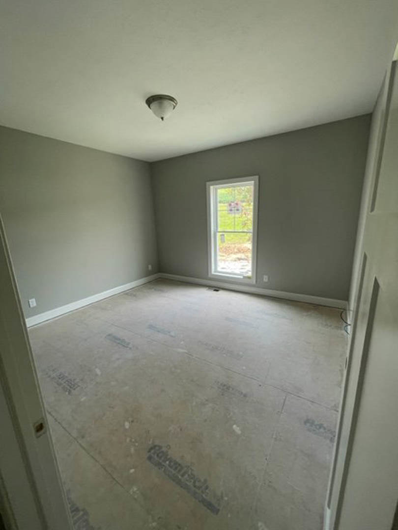 Concrete floor and plaster walls in an unfinished room with a large window overlooking a green lawn, white pipe visible near the wall.