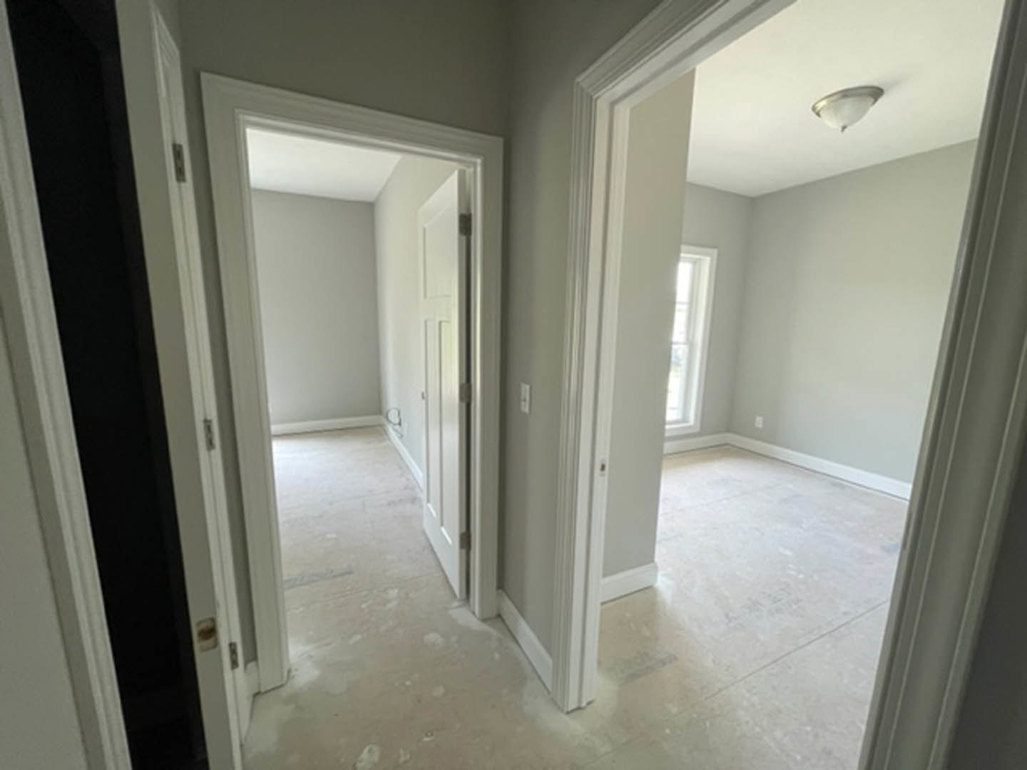 Hallway with smooth white walls, white door and door frame, ceiling light fixture, partial view of window, light-colored flooring, simple molding