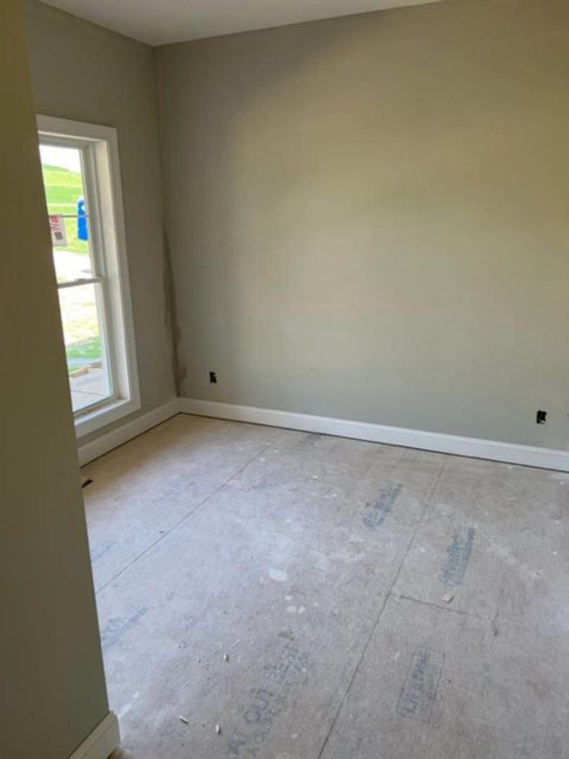 Bright room with white plaster walls, large window overlooking green lawn, unfinished concrete floor with scattered wood planks