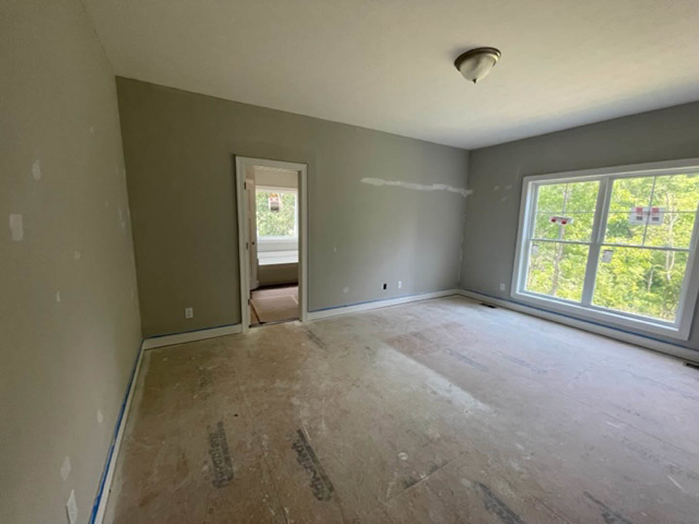 White plaster walls and ceiling, large window with trees visible outside, wood door with sign, dirty light-colored flooring, close-up of modern light fixture