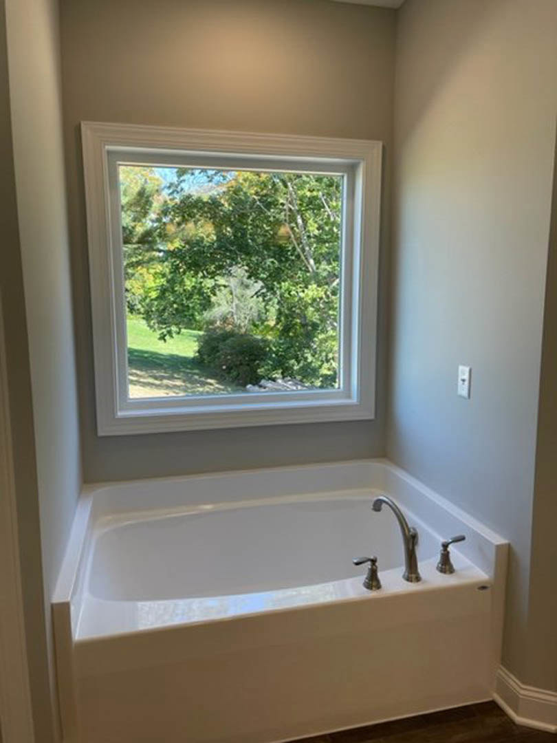 Freestanding bathtub beneath large window overlooking leafy trees, chrome faucet, light-colored walls, modern bathroom interior