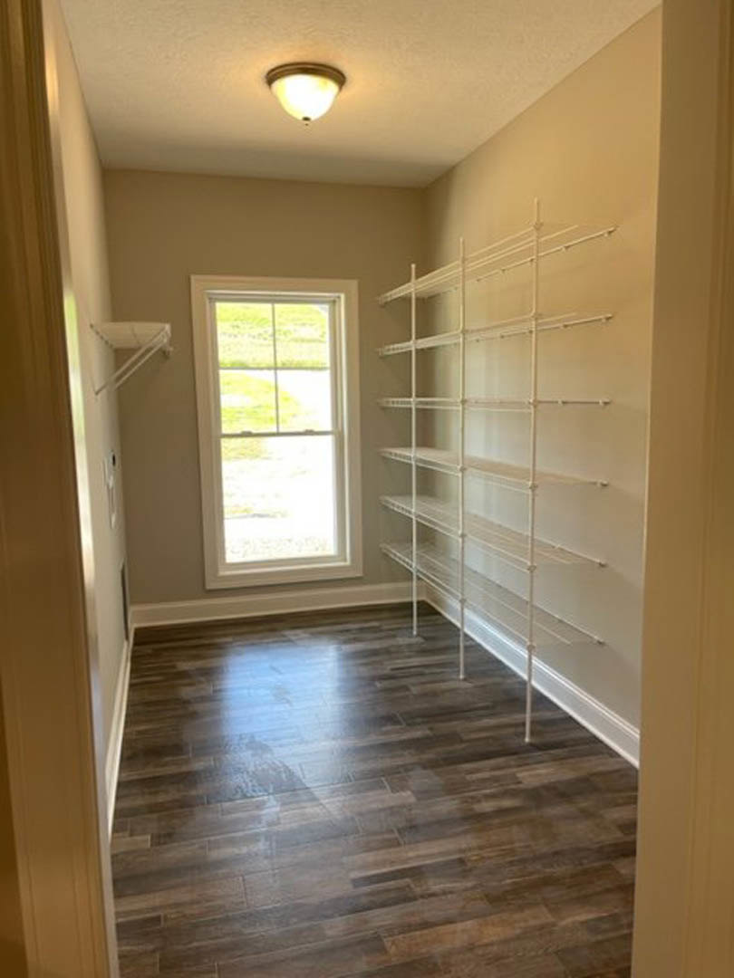 Built-in white shelving along one wall, large window overlooking grassy yard, hardwood flooring with glossy finish, white stair railings, ceiling-mounted light fixture.