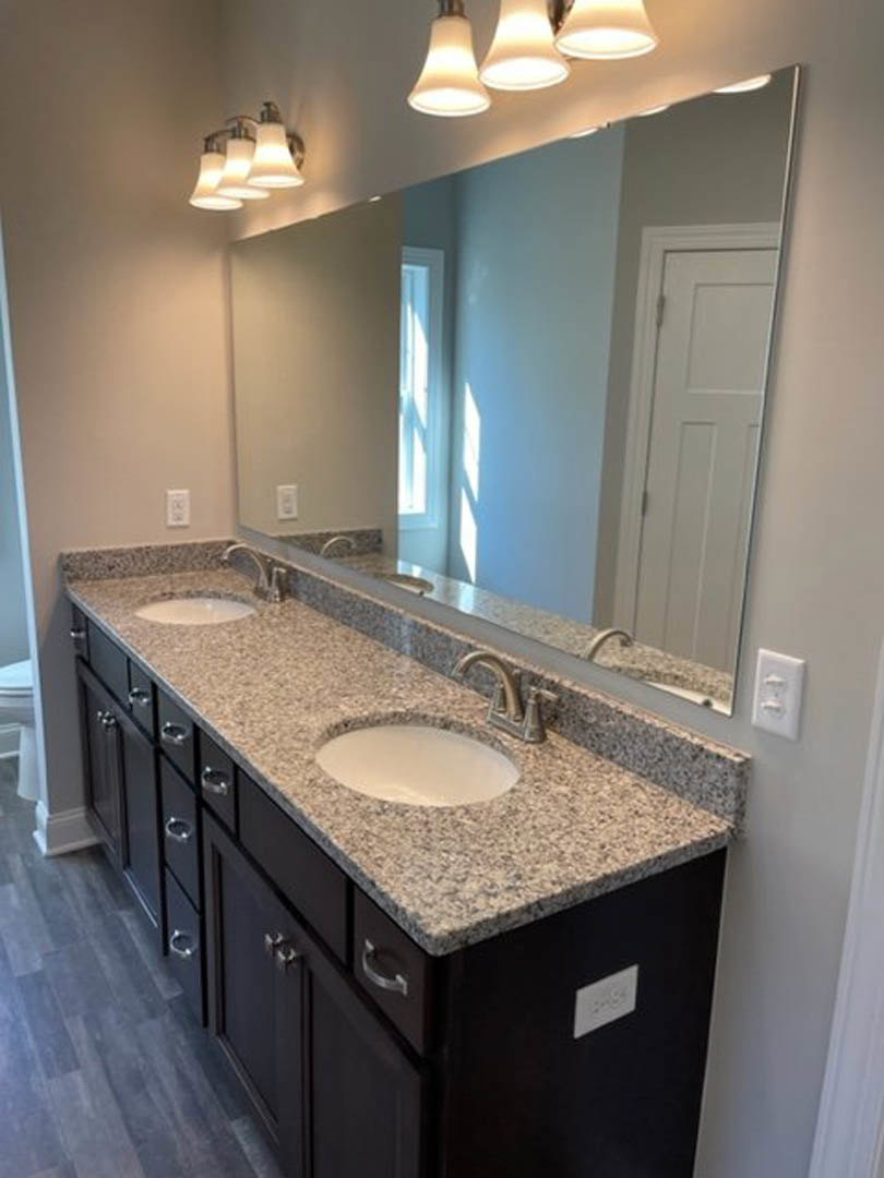 Spacious bathroom featuring a wide wall mirror above a dark vanity with white countertop, modern ceiling light fixtures, wood flooring, and tiled walls.