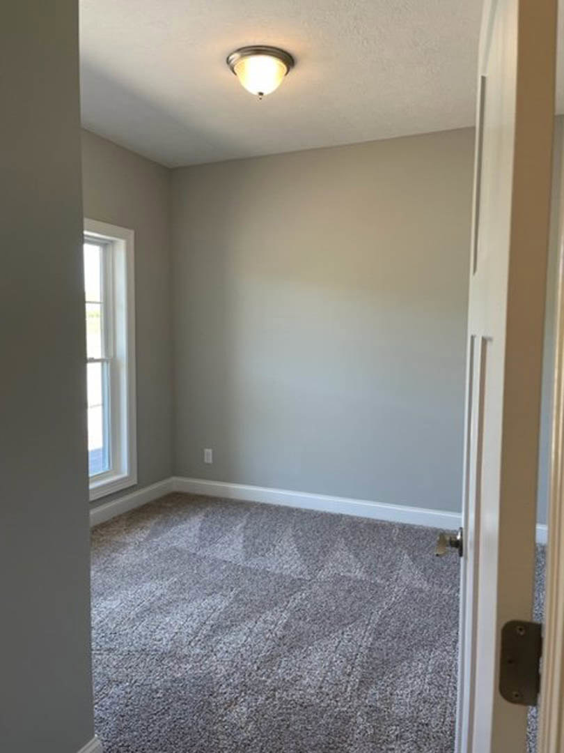 Neutral-toned carpeted room with white walls, ceiling-mounted light fixture, and window framed in white trim