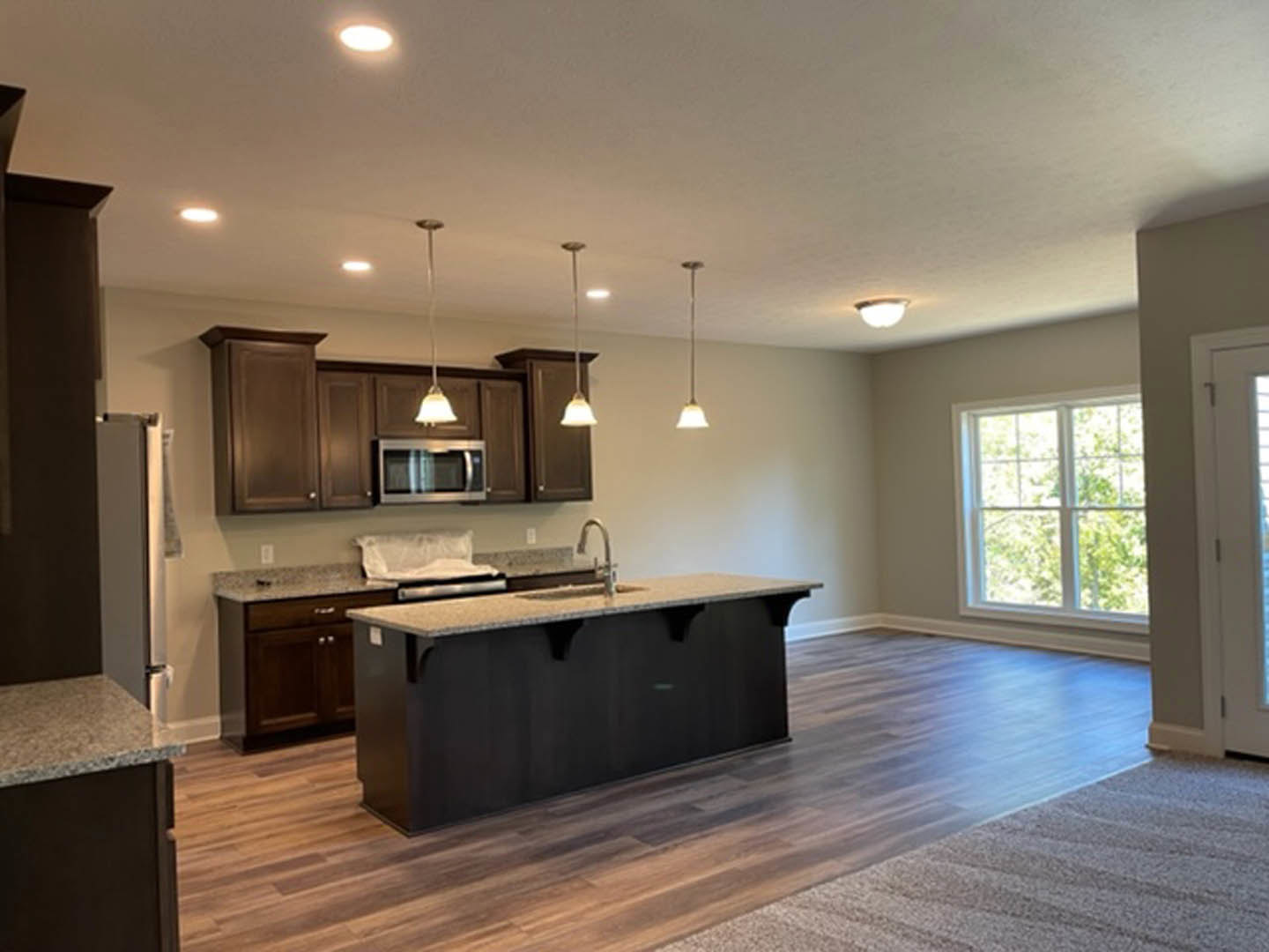 Spacious kitchen featuring a large central island with stone countertop, wood plank flooring, white cabinetry, stainless steel sink, built-in microwave, and a window overlooking