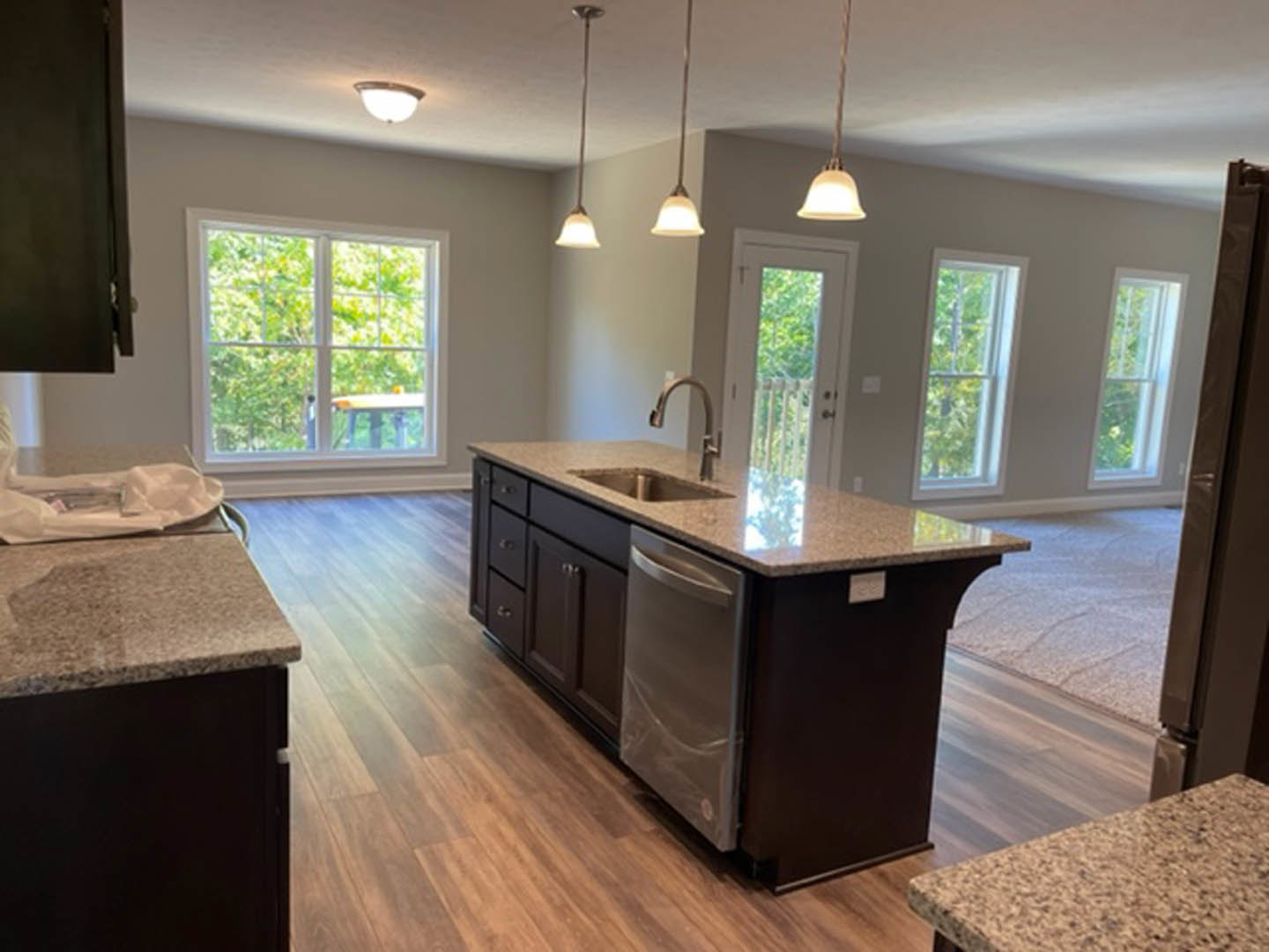 Open kitchen with light wood flooring, white cabinetry, stone countertop, large kitchen island featuring dual sinks, window overlooking green trees, stainless steel appliances, and
