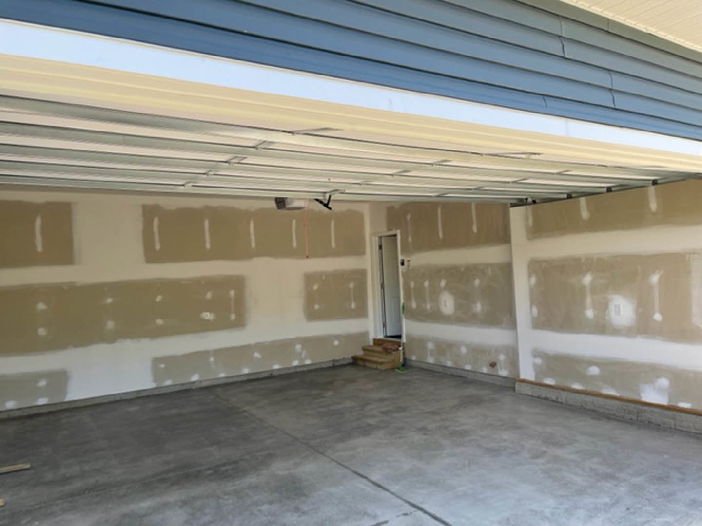 White-painted garage wall, exposed ceiling beams, concrete floor, adjacent door, empty interior space