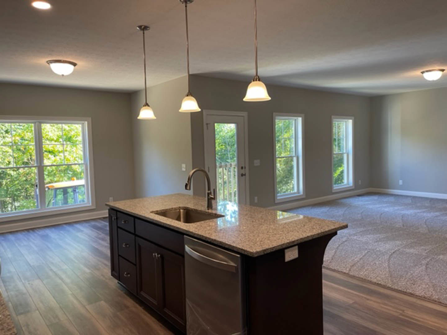 Open kitchen and living room featuring light wood cabinetry, stone countertops, stainless steel dishwasher, undermount sink with chrome faucet, large windows overlooking trees