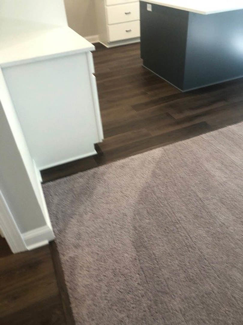 White cabinet with silver handles beside a brown rug on hardwood floor, white shelf with brown base, corner of kitchen counter visible.