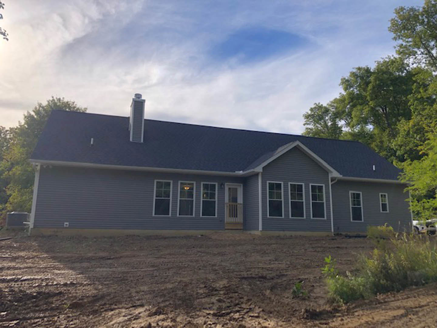 Two-story house with beige siding, covered front porch, brick chimney, multiple windows, and balcony; dirt yard in foreground, sparse landscaping, cloudy sky overhead