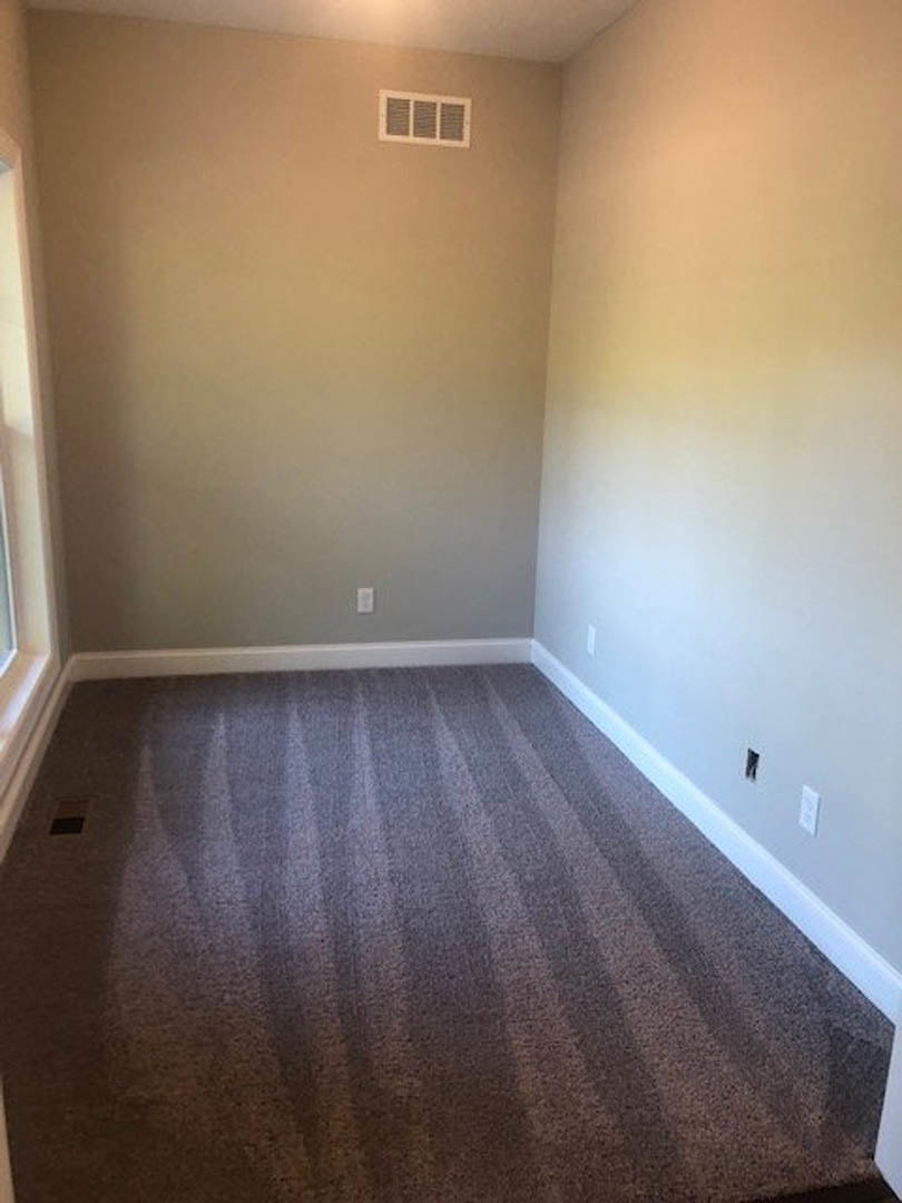 Striped carpeted floor in a room with a large window, white plaster walls, wall and floor vents, and laminate wood trim