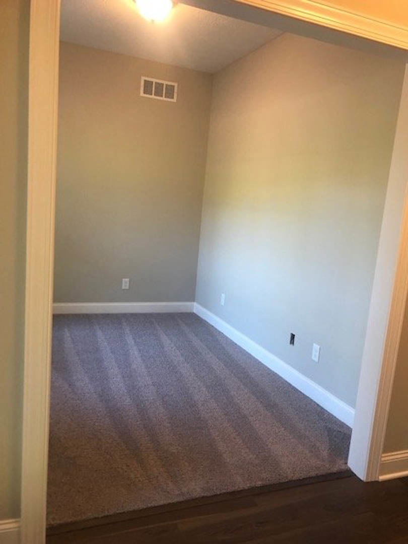 Carpeted floor with striped pattern, white walls, ceiling light fixture, air vent, and wood trim around doorway